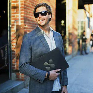 Man smiling, holding laptop, wearing sunglasses and a blazer.